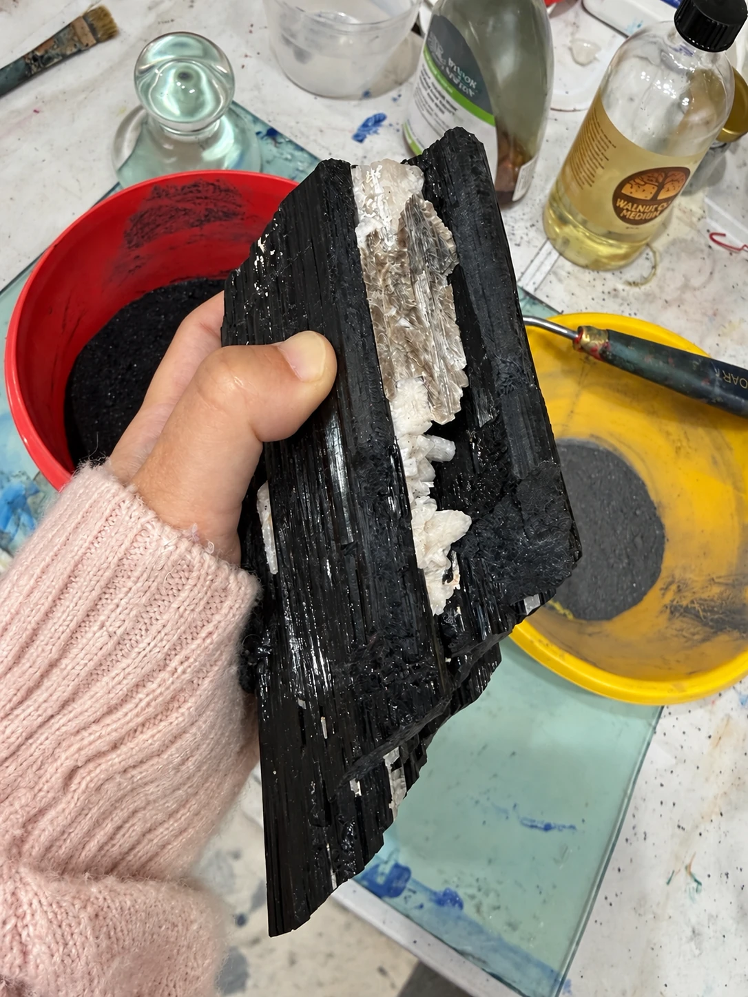 A hand holding a dark mineral with a white vein, in front of containers of pigments.