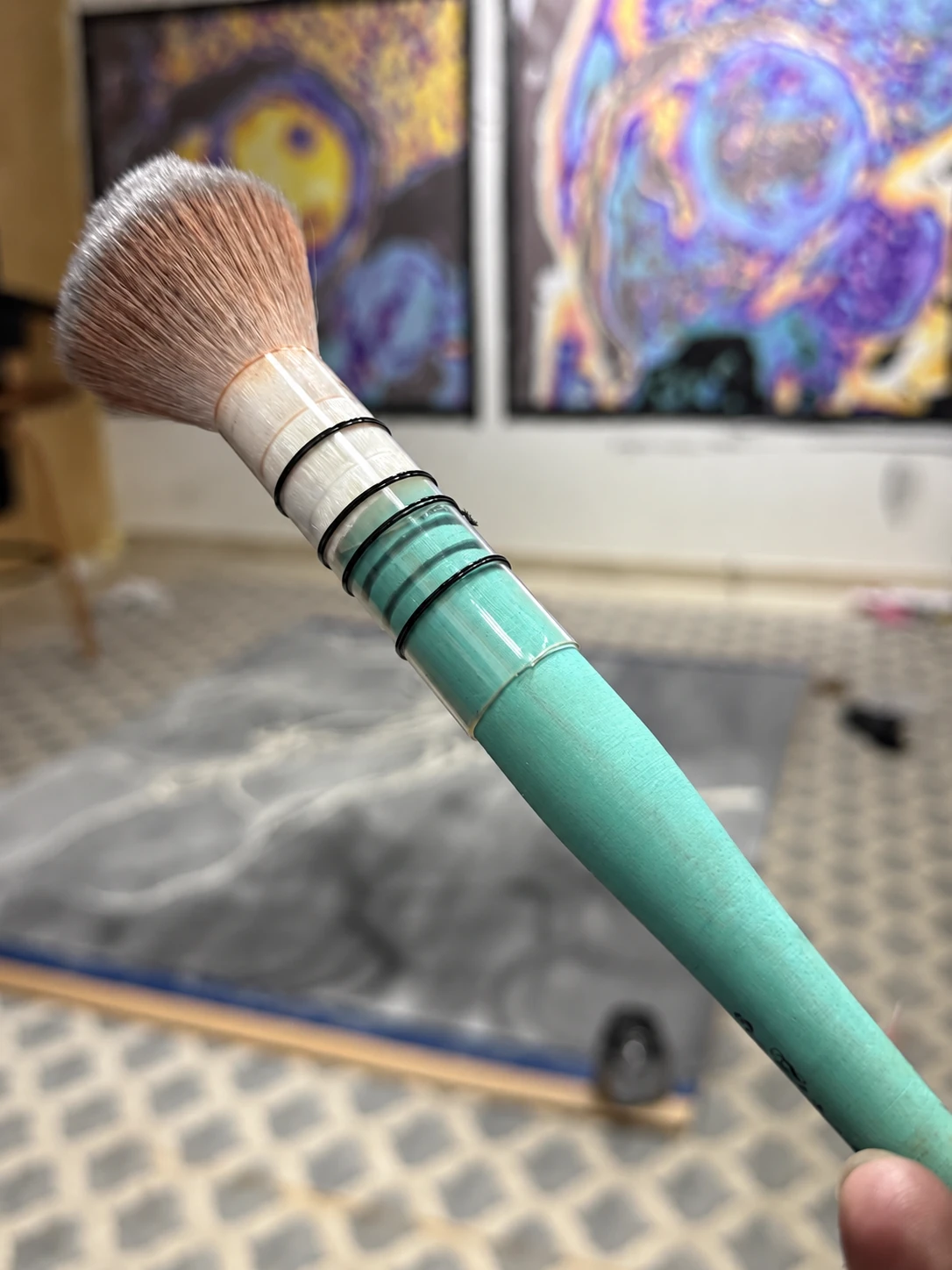 A hand holding a brush with a turquoise handle, with artworks in the background.