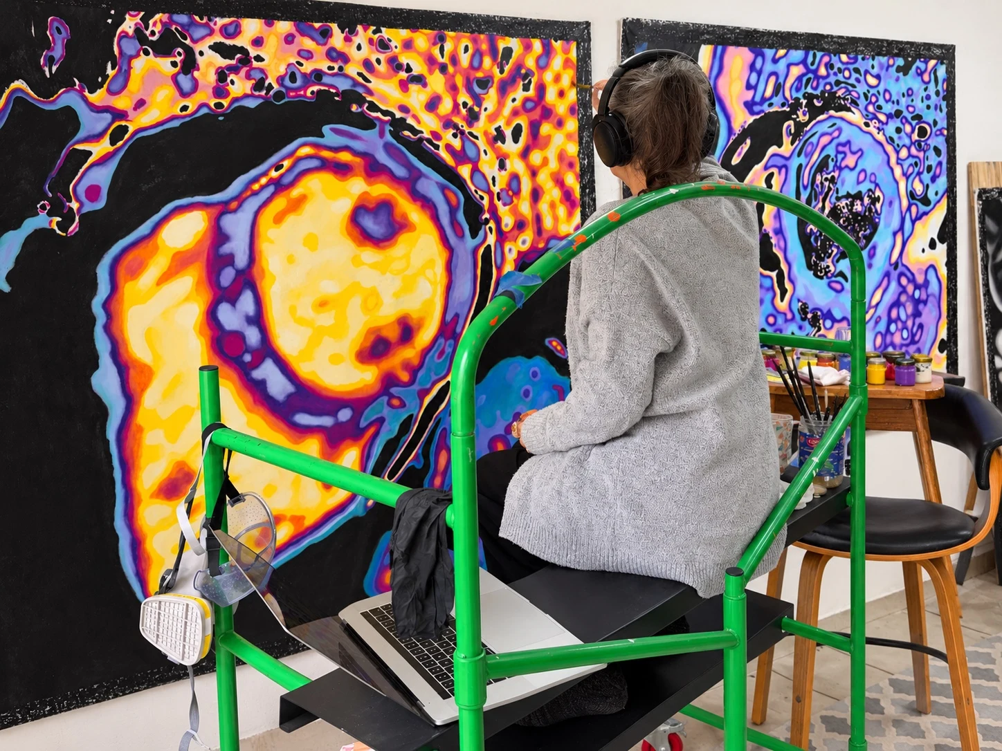 Boo Shvartsman seen from behind, sitting on a green bench in the studio while painting two colorful canvases.